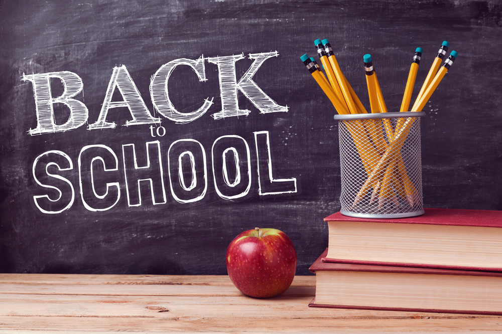 Chalk block letters saying back to school with a container of pencils on two red books and a red apple on the table